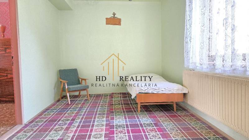 Lôžko a kreslo v miestnosti rodinného domu s farebným kobercom a záclonou.